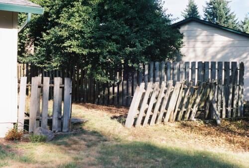 A dilapidated wooden fence surrounds a grassy area, part of it leaning. 