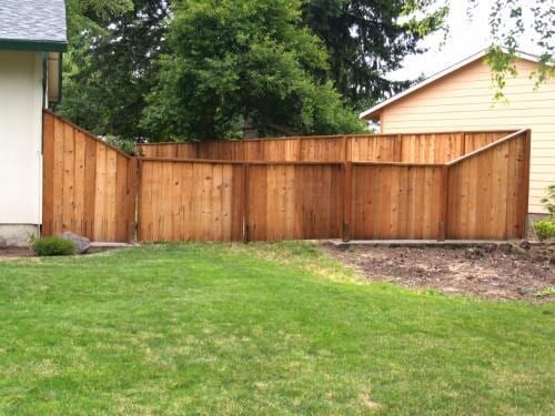 Wooden fence around a grassy backyard, adjacent to two houses and trees.