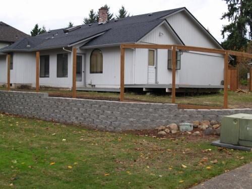 Backyard with a retaining wall, lawn, and house with a wooden pergola structure.