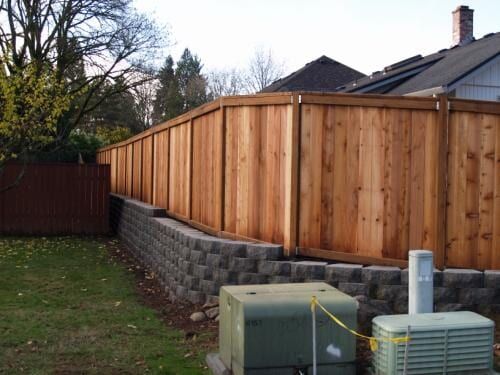 Wooden fence on top of a retaining wall in a backyard with electrical boxes in the foreground.