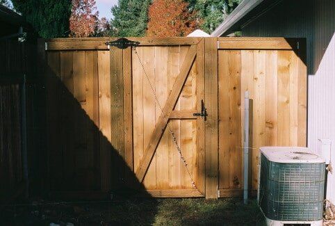 Wooden gate in a backyard, with a metal handle, diagonal support, and an air conditioning unit beside it.