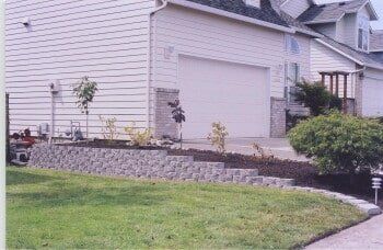 Stone retaining wall in front yard, tiered with landscaping, and a garage behind it.