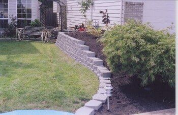 Stone retaining wall in a yard, with grass lawn and a bush, near a house with a window.