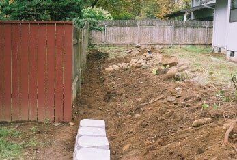 Dirt trench next to a red fence and house, with retaining blocks and soil.