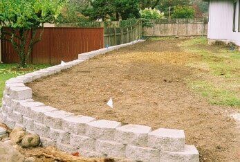 Retaining wall made of gray blocks surrounds a dirt yard, with brown fence and house in background.
