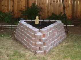 Triangular brick planter filled with dark soil, Buddha statue, set in a grassy backyard, wooden fence in the background.
