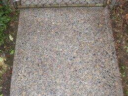 Concrete sidewalk, textured with exposed aggregate, next to greenery, leading to a metal gate.