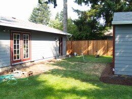 A backyard with a house, wooden fence, and grass. The house has red-framed doors.