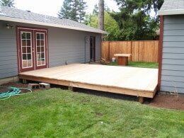 Wooden deck attached to a house with double doors, on a grassy yard, with a fence in the background.