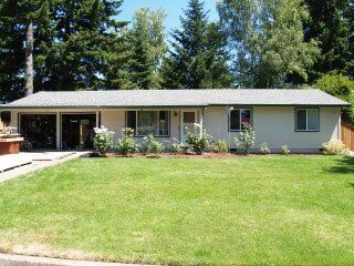Ranch-style house with garage, beige siding, green lawn, rose bushes, and tall trees in the background.