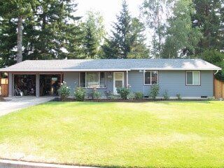 Blue ranch-style house with attached carport and garage, green lawn, and trees in the background.