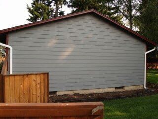Gray-sided building with brown trim and white gutters, next to a brown wooden fence, set in a yard.