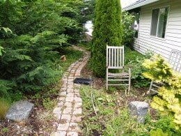 Brick path leads through a garden with a chair beside a house.