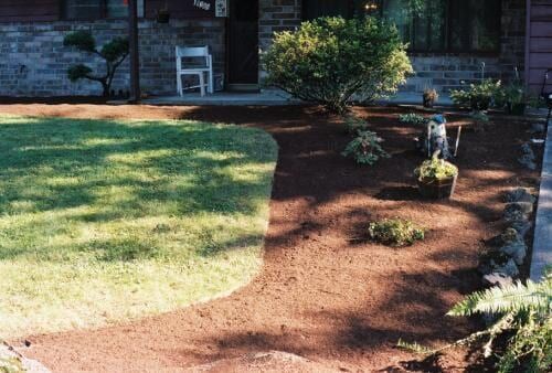 Lawn and landscaped garden in front of a brick house with a white chair on the porch.