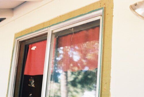 Close-up of a window with a red curtain inside, framed by a beige and green border, set against a white wall.
