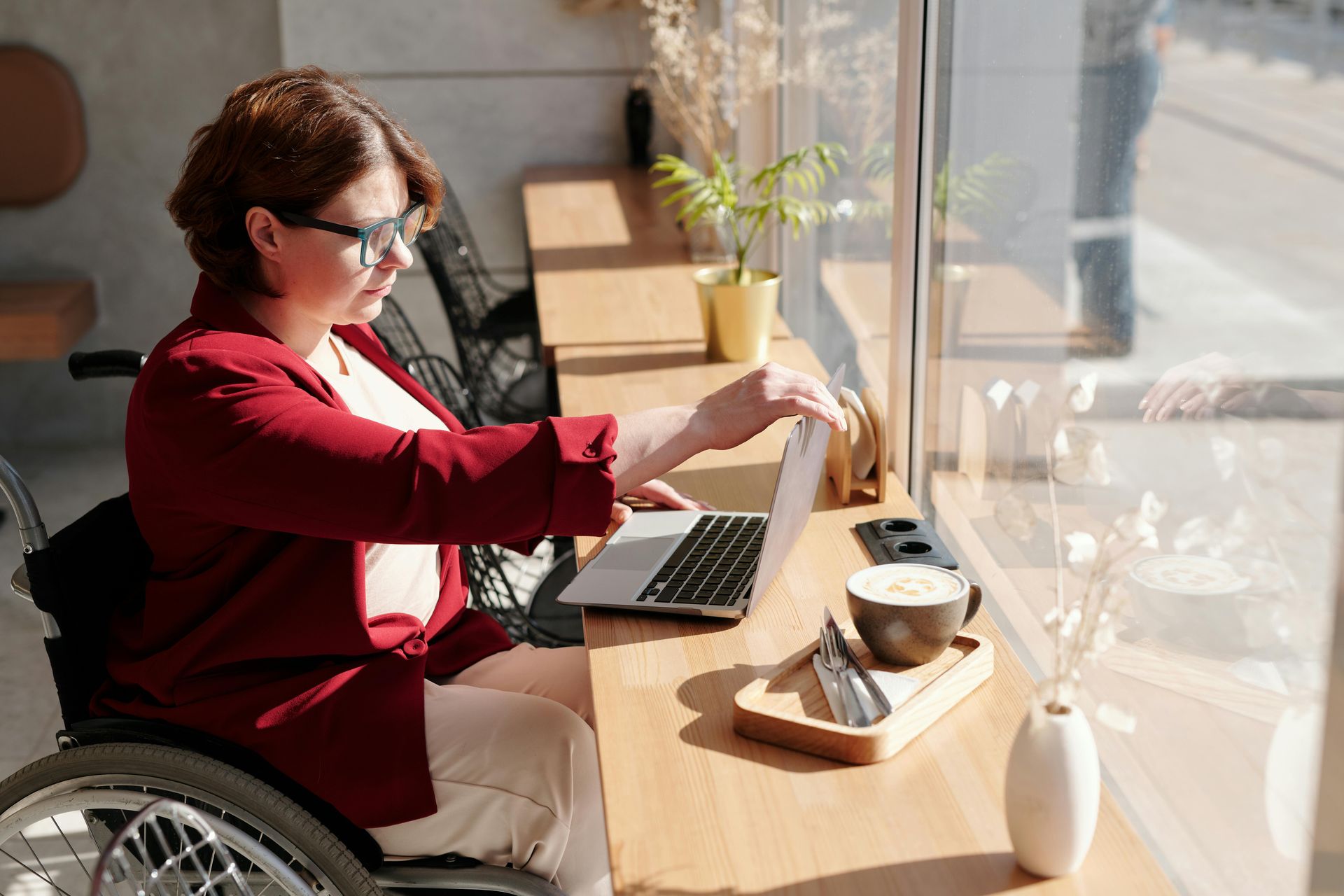 Woman in wheelchair using laptop at a cafe table near a sunny window.