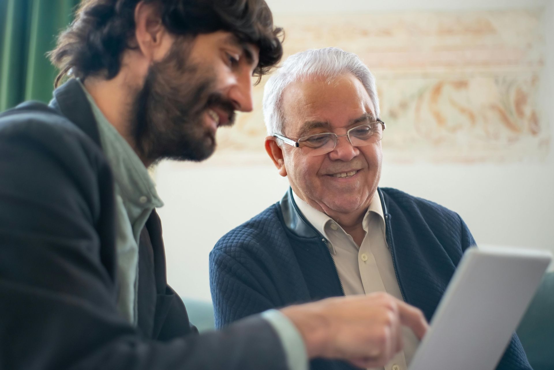 Man in a suit jacket shows a tablet to an older man, both smiling. Indoors, near a decorated wall.