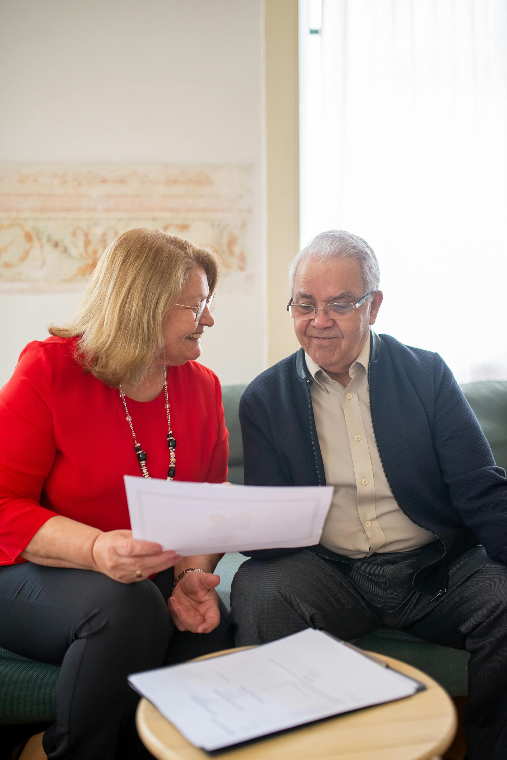Woman in red and man in blue cardigan looking at documents together, seated in a living room.