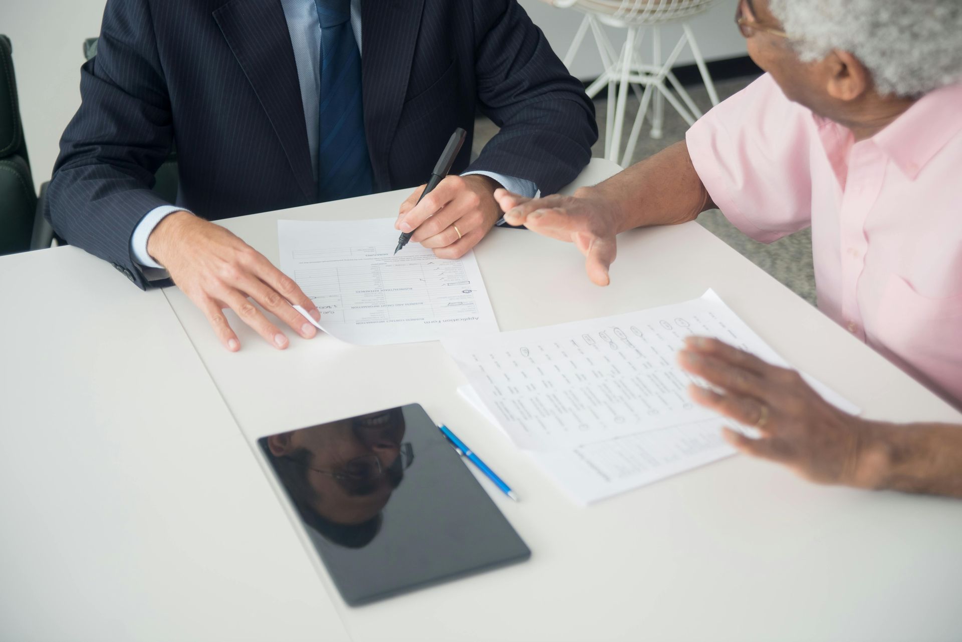 Man in suit signing documents, woman gesturing at the table, tablet, and pen on a white table.