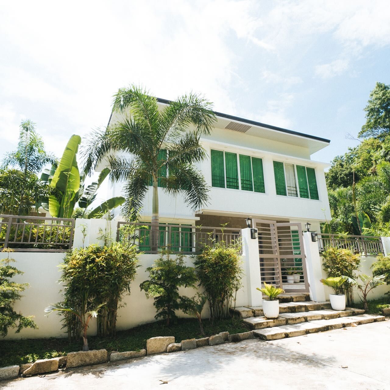 White two-story house with green shutters, palm trees, and a garden against a bright sky.
