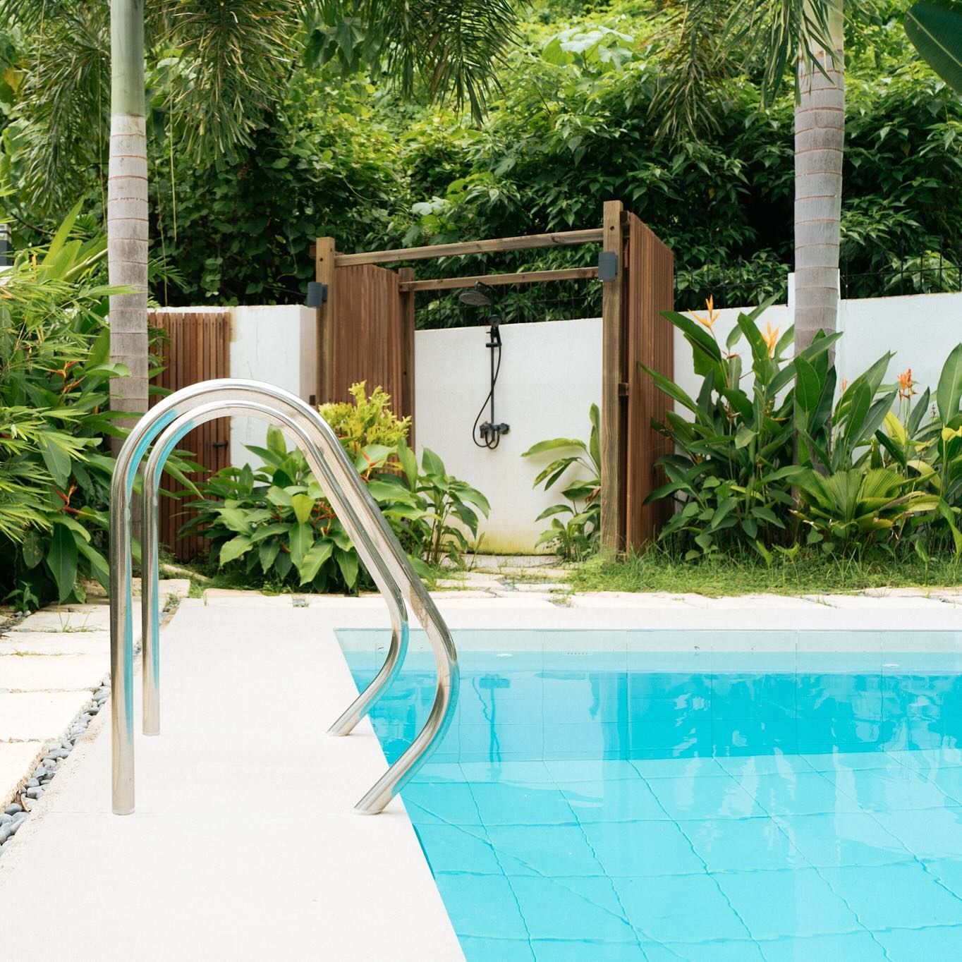 Pool with a metal ladder. Wooden outdoor shower in the background surrounded by lush green plants and trees.