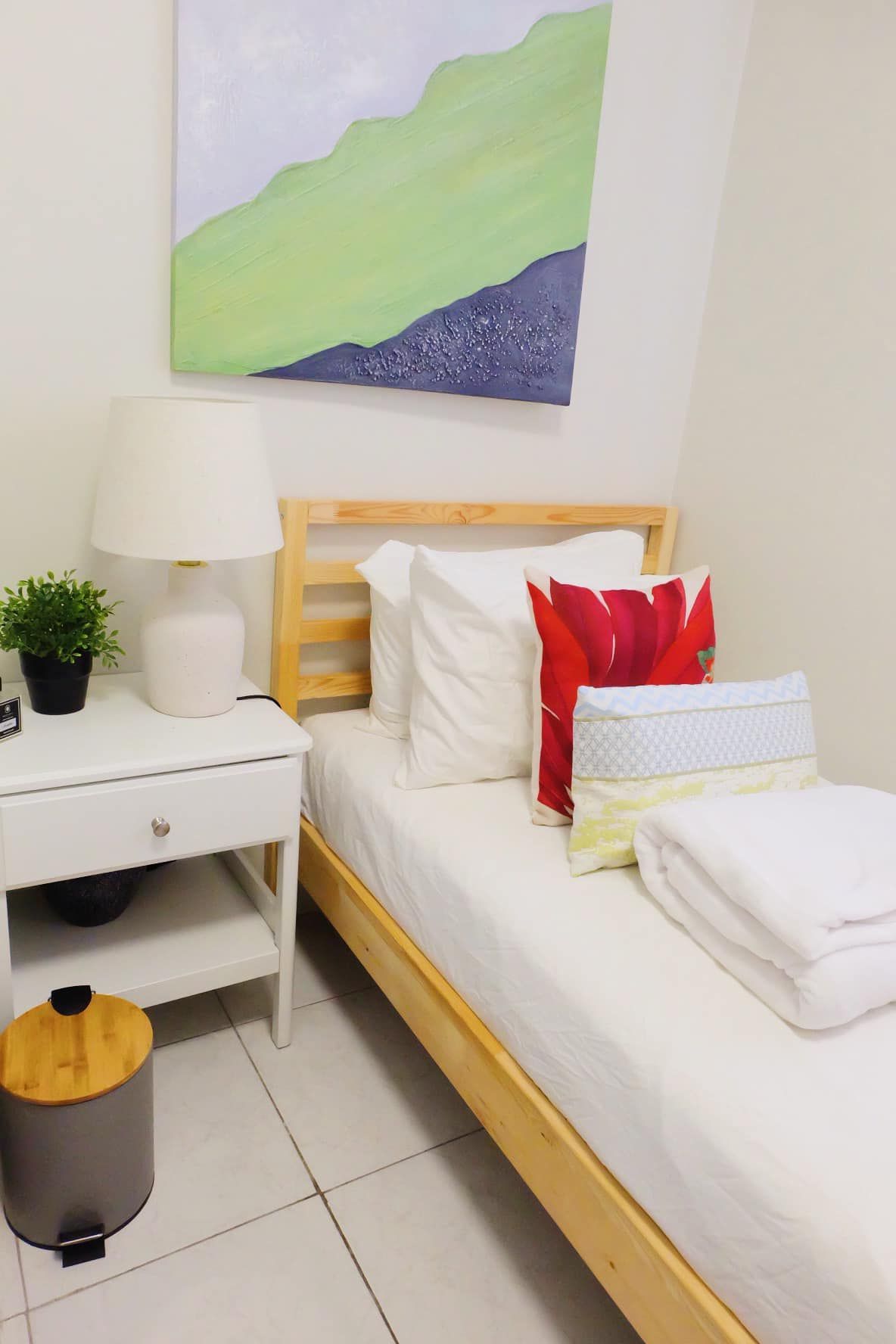 Bedroom with a single bed, nightstand, and colorful pillows. Artwork on the wall, gray trash can.