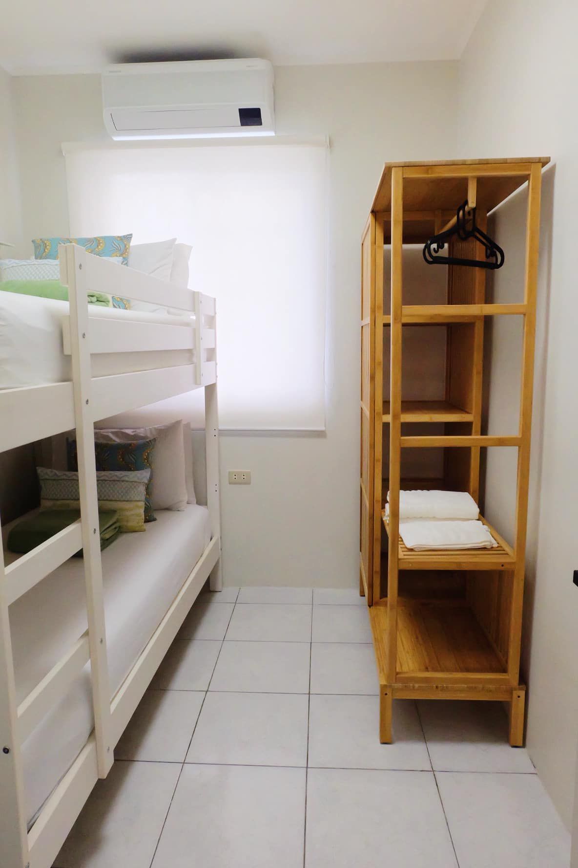 A small, white-walled bedroom with bunk beds, a wooden open wardrobe, and a window with a blind.