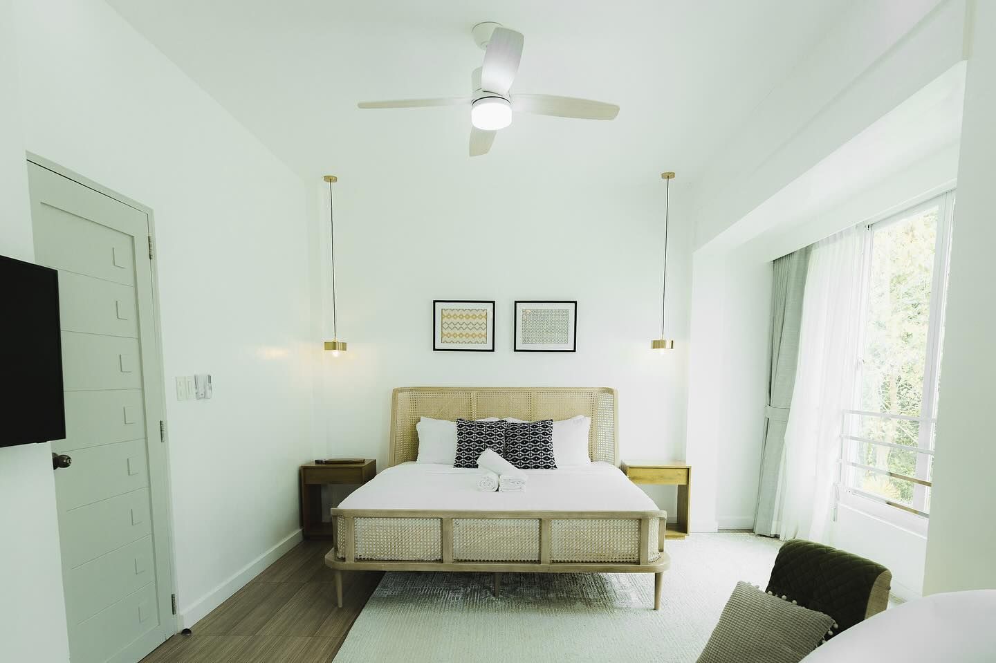 Bright bedroom with a light wood bed, two bedside tables, and a large window.  Two hanging lights, ceiling fan.