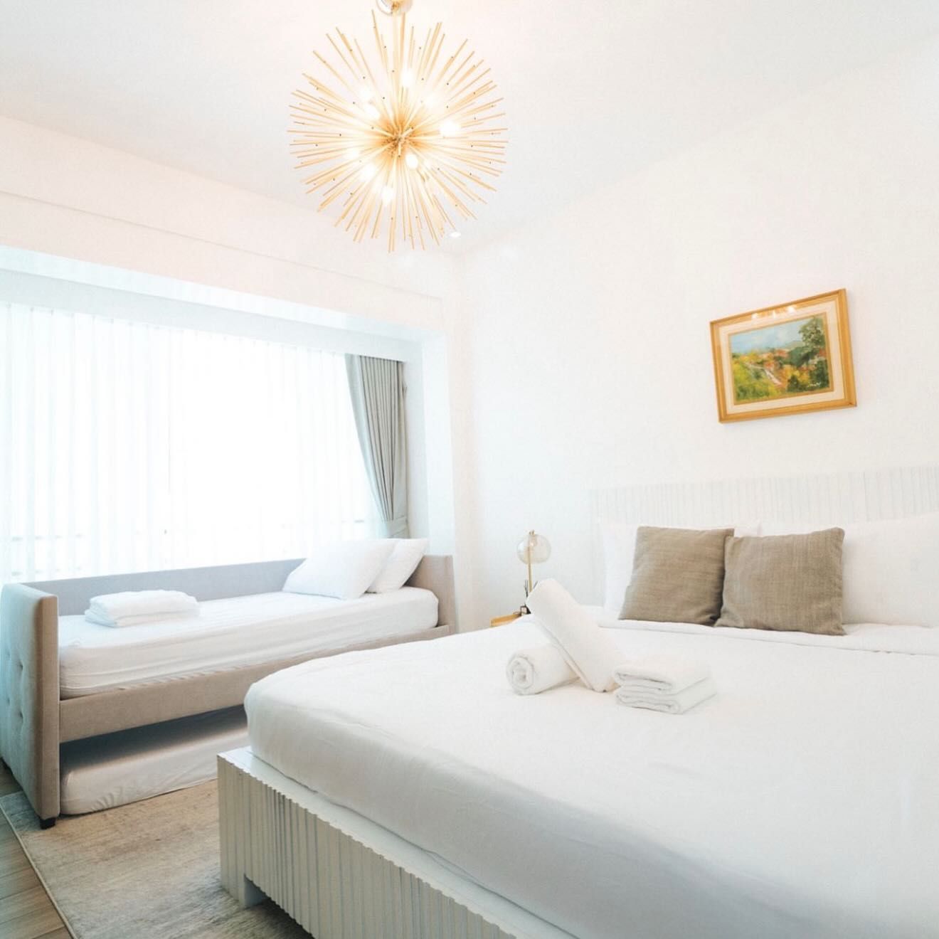Bright bedroom with white bed, gray sofa, painting, and starburst chandelier.