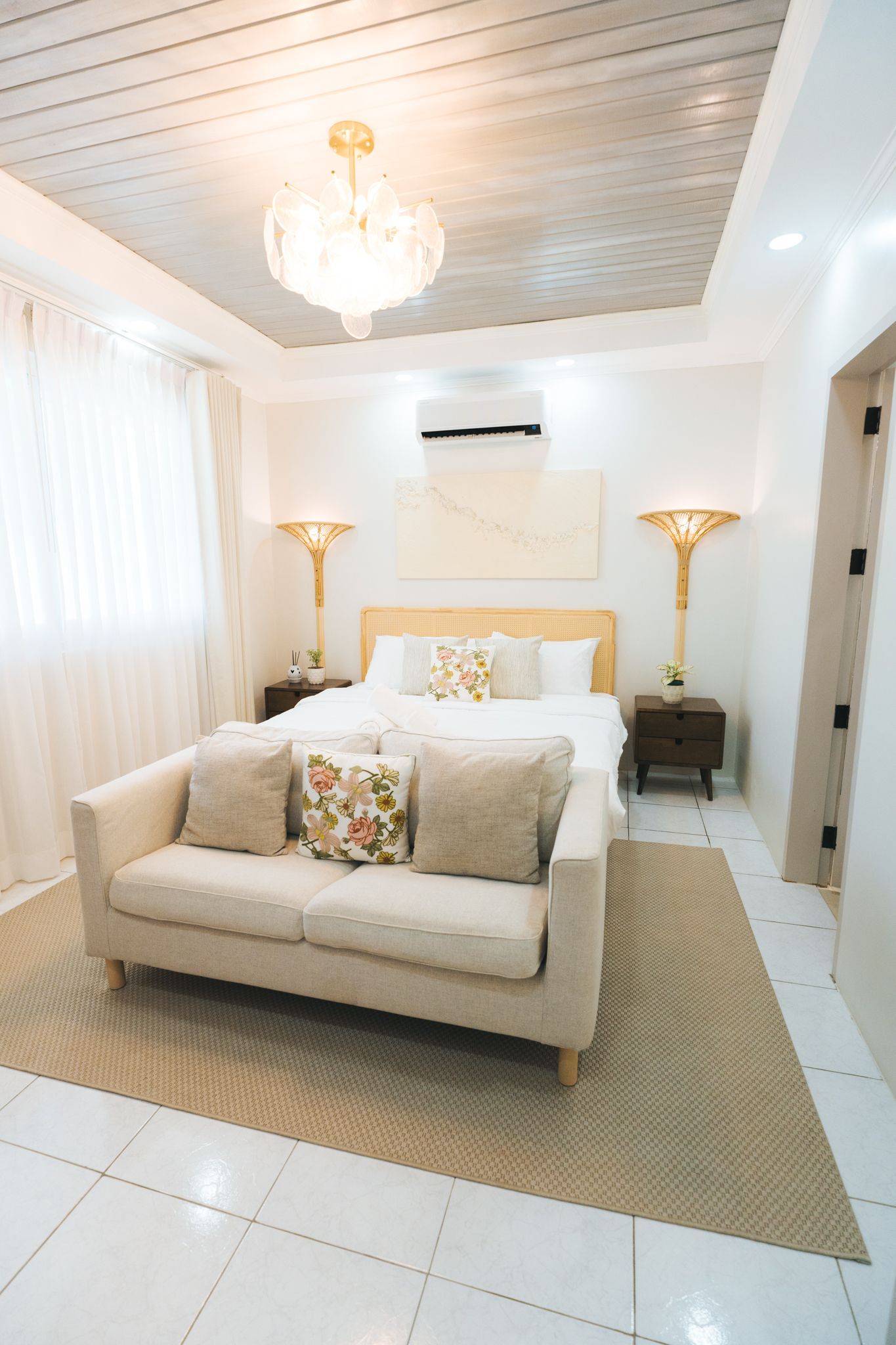 Cozy white bedroom with a bed, sofa, chandelier, and lamps; a neutral rug and light floors.