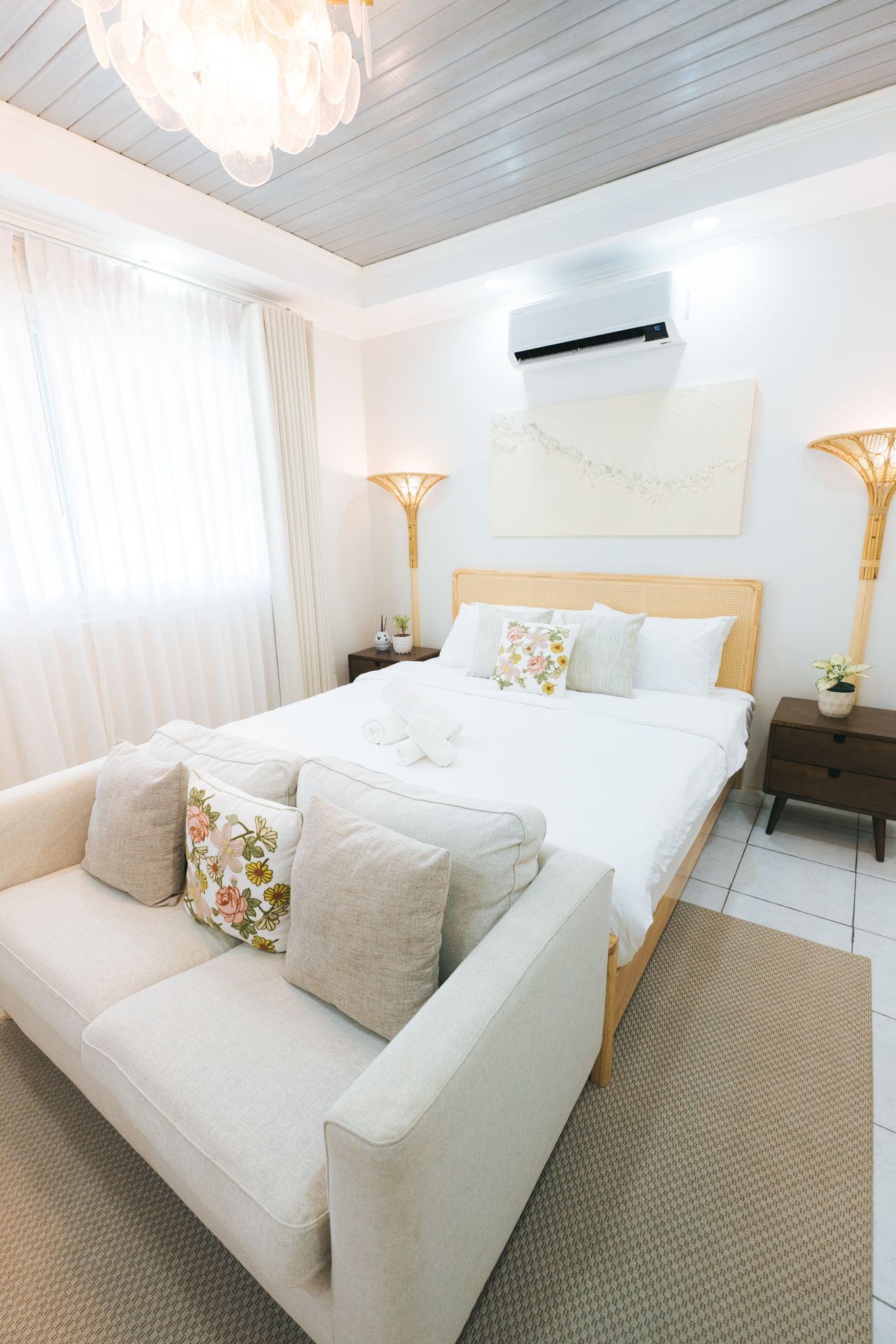 Bright bedroom with a sofa, bed, lamps, and a decorative wall piece; white and neutral tones.
