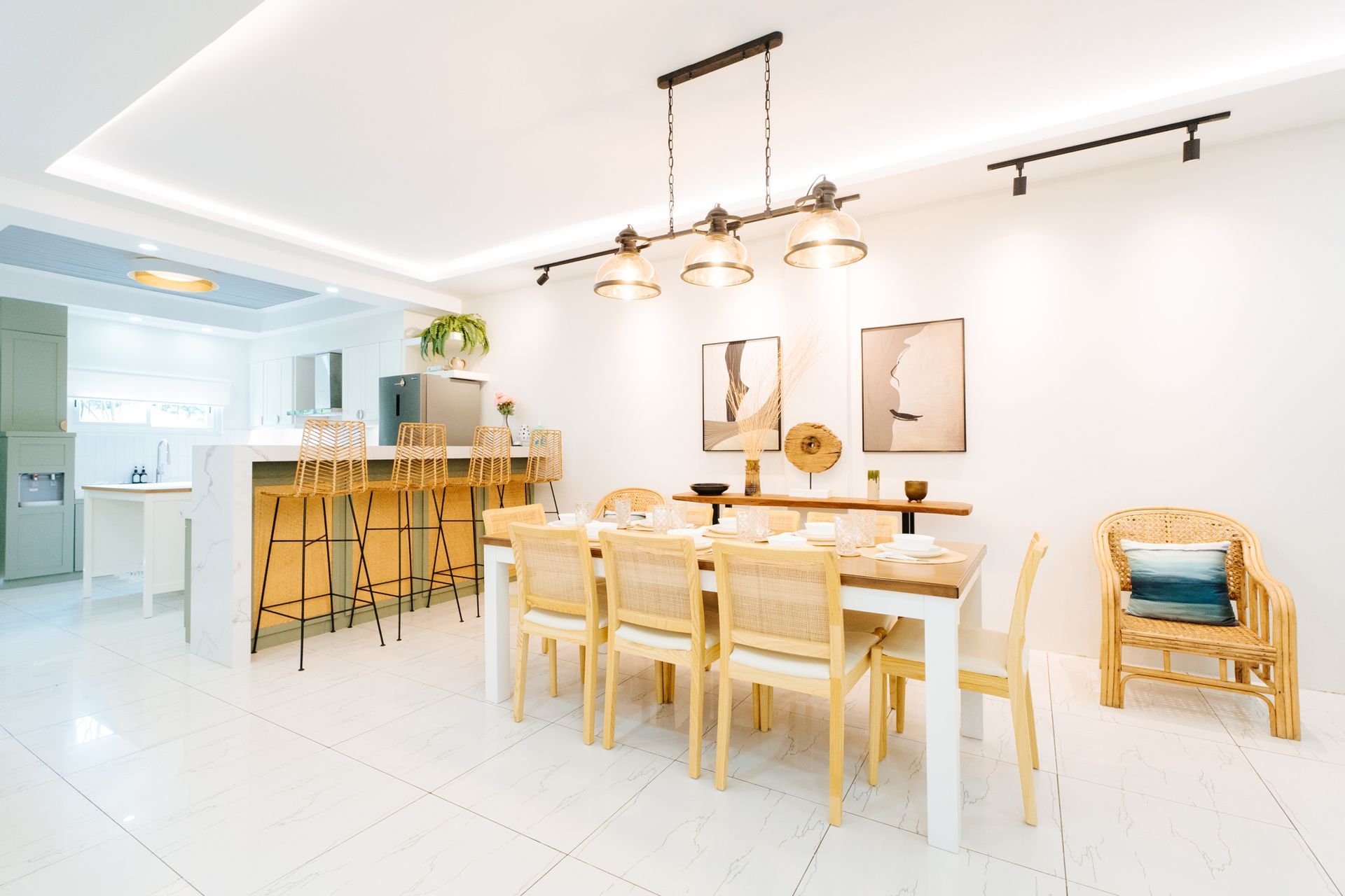 Bright, modern dining room with a long table, chairs, and overhead lighting. Kitchen bar and artwork visible.
