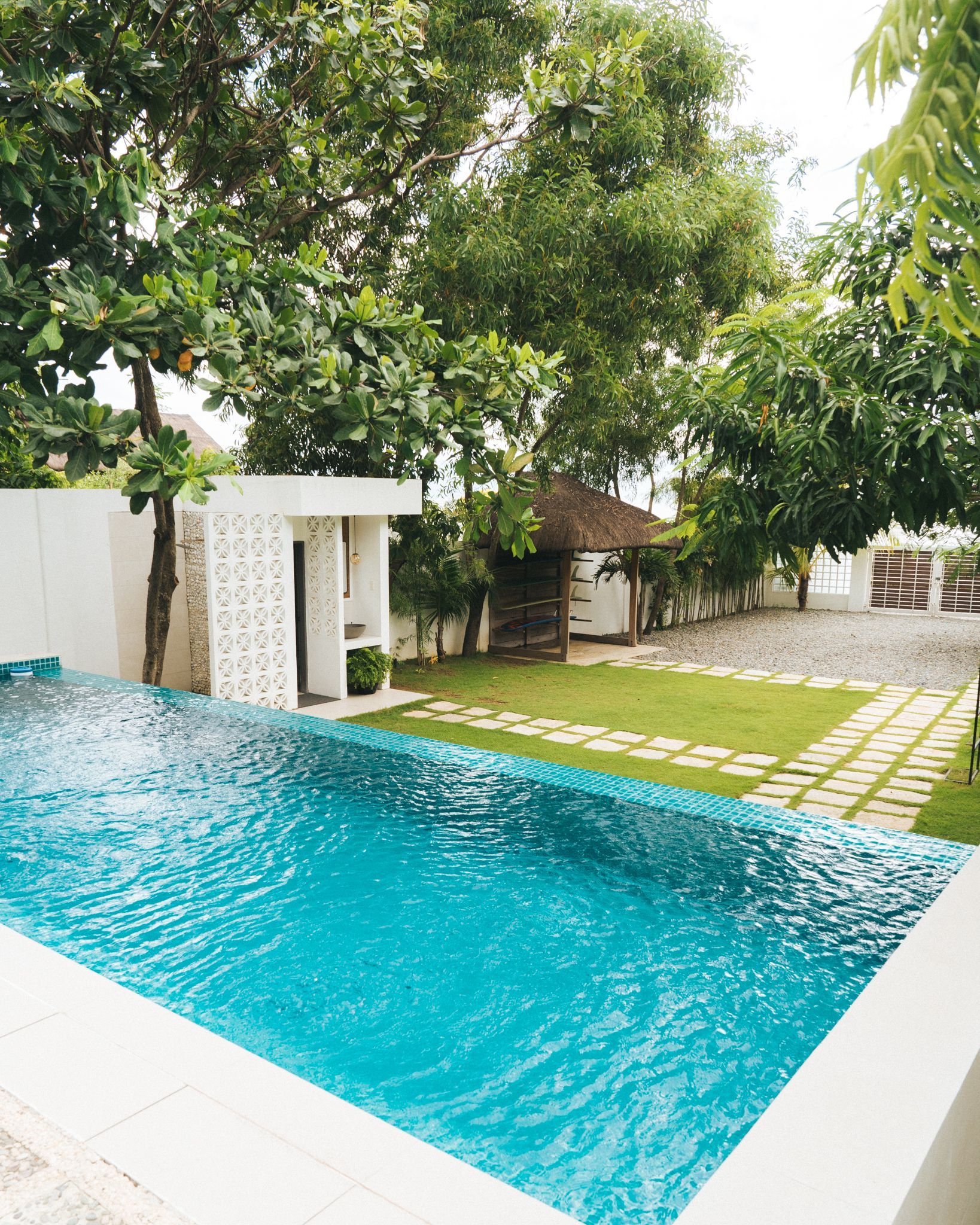 Swimming pool next to white wall, surrounded by green lawn and trees. A small hut sits in the background.