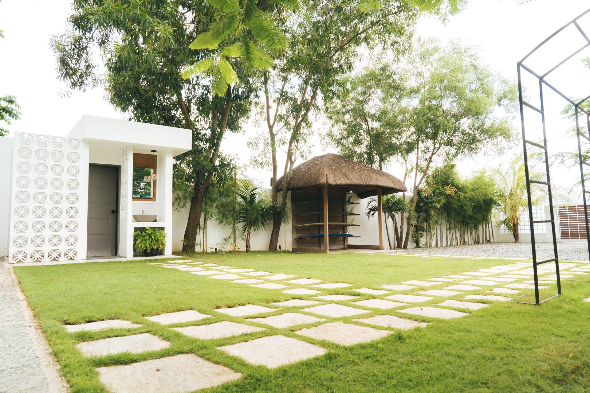 Green lawn with stone pathway, white building with grill, wooden gazebo, and trees.