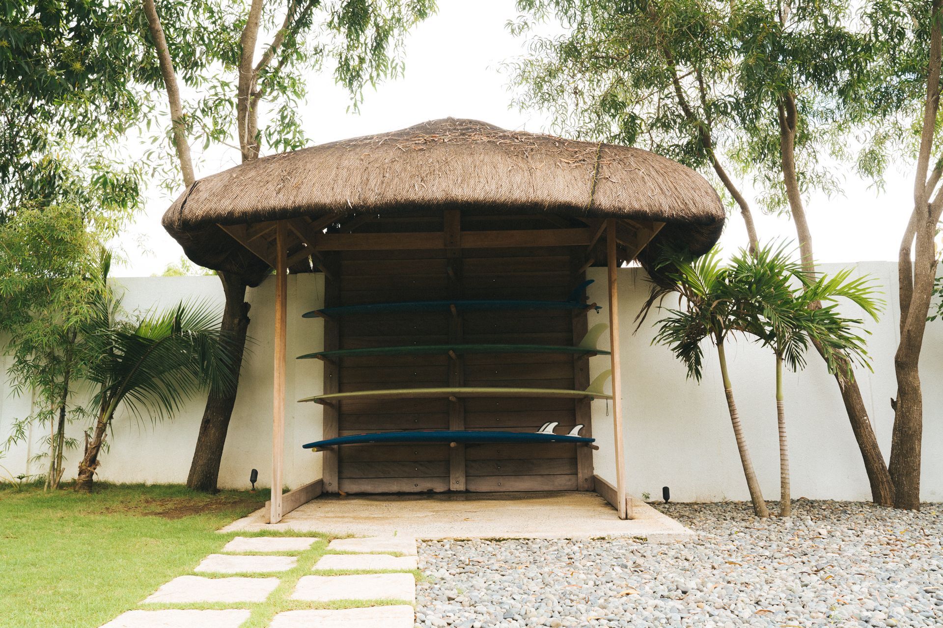 A thatched-roof structure with surfboard storage, set in a yard with a stone path and white wall.