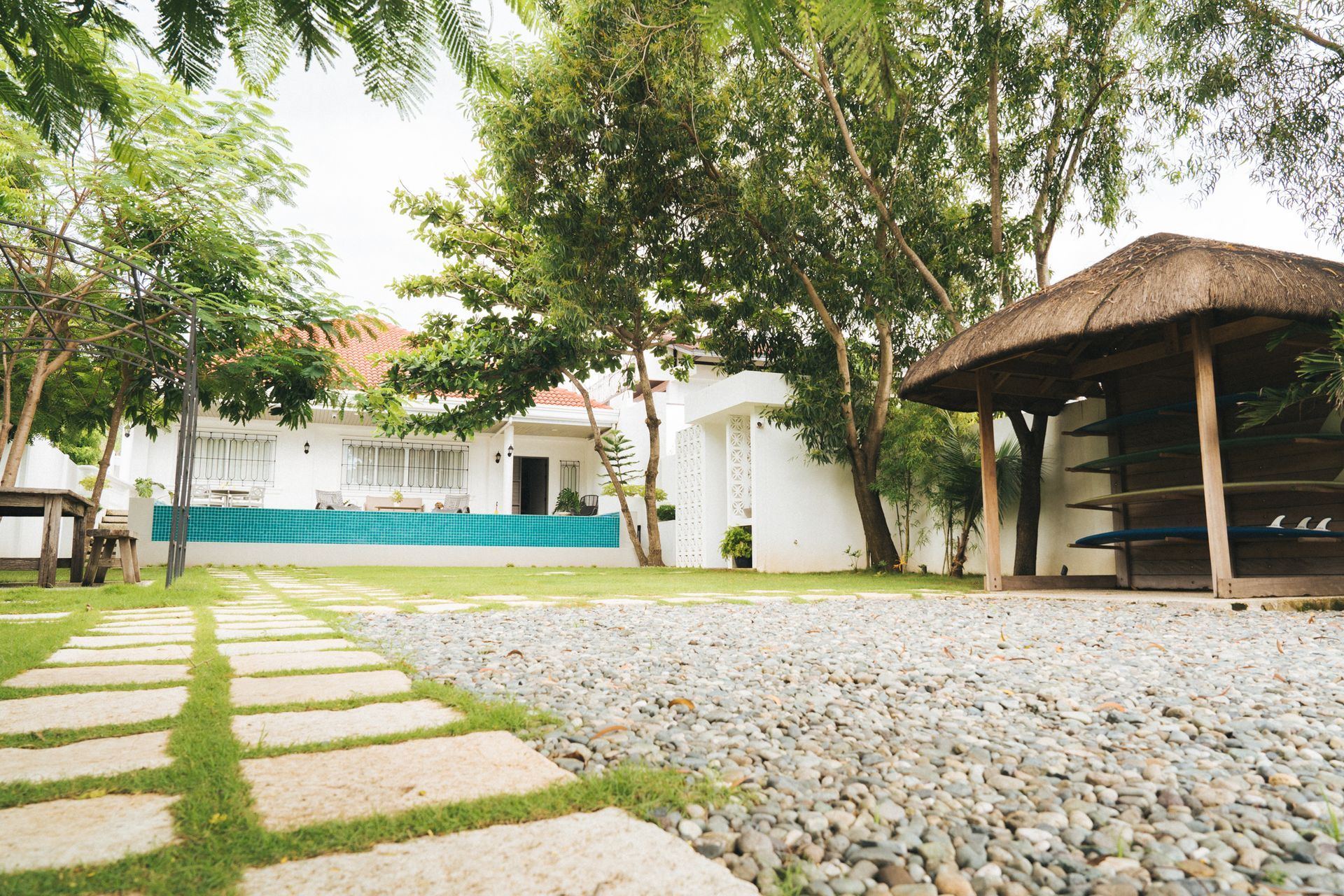 Stone path leads to white building with teal accent, surrounded by trees and thatched-roof gazebo.