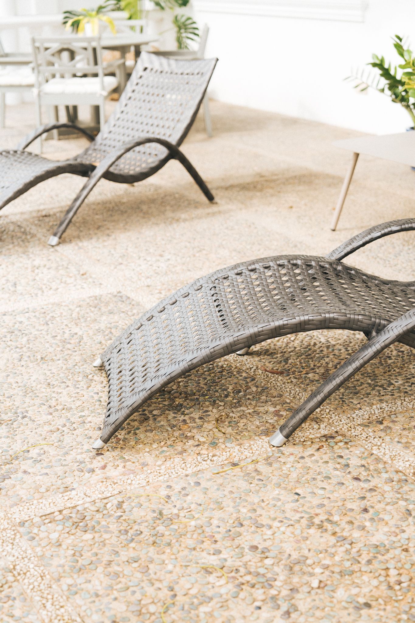 Two gray wicker chaise lounges on a pebbled patio, with a table and chairs in the background.