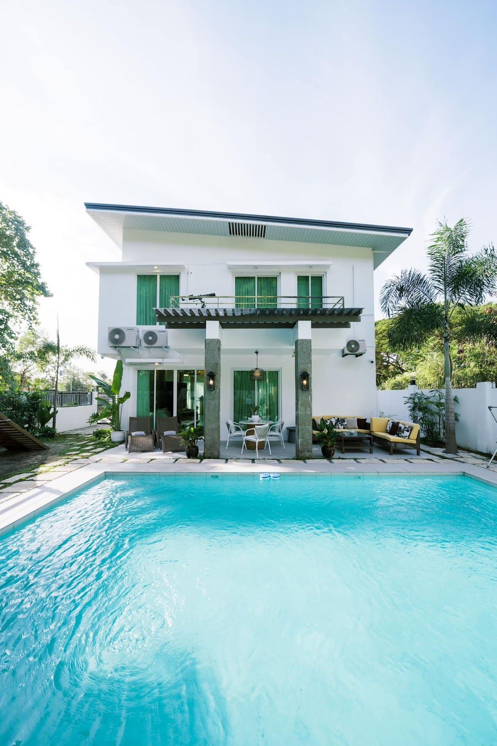 Two-story white house with pool; turquoise water. Green window frames, outdoor furniture, sunny day.