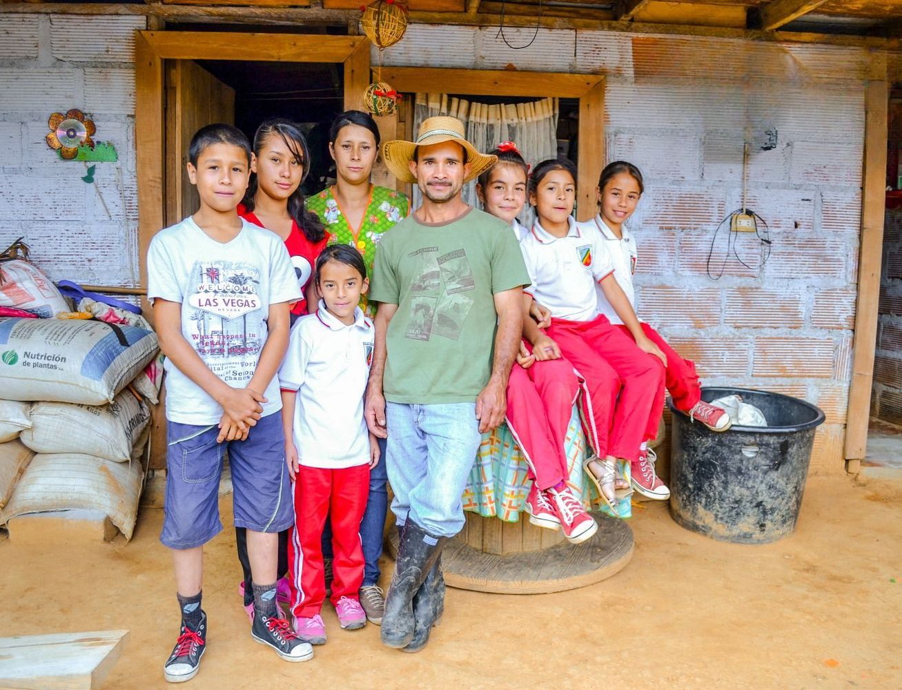 A family of nine stands in front of their home; father in hat and boots, children in school uniforms, setting is a rural area.