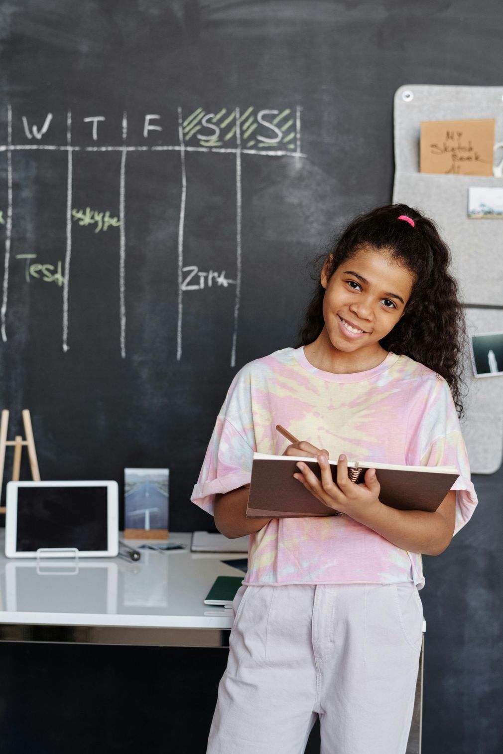 A smiling Black girl with curly hair, holding a notebook, stands in front of a chalkboard schedule and desk.