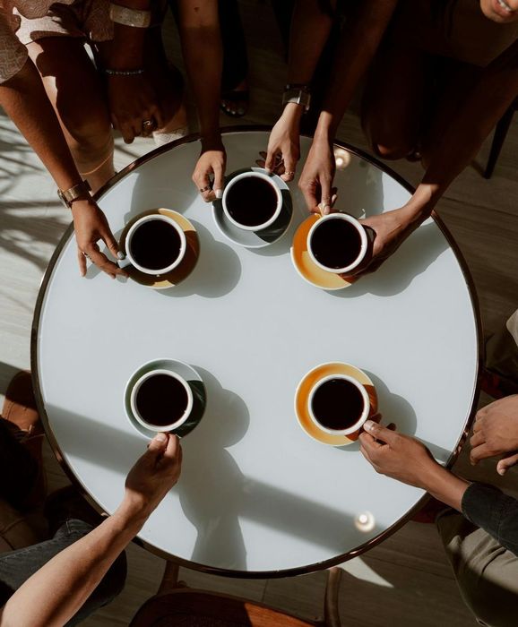 People at a round table, reaching for coffee cups. Overhead view with hands and colorful saucers.