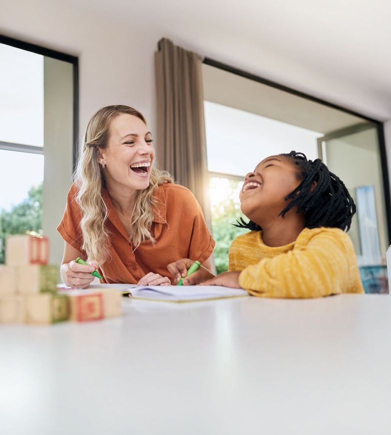 Woman and girl laughing together at a table, working on a notebook with wooden blocks nearby.
