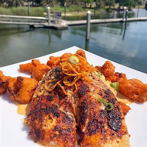 A close-up of a seared, seasoned fish fillet topped with crispy fried onions, served with sides, overlooking a lake.