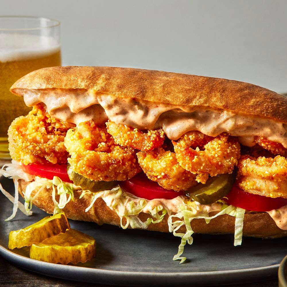 A fried shrimp po' boy sandwich with remoulade sauce, tomatoes, pickles, and lettuce on a plate next to a glass of beer.