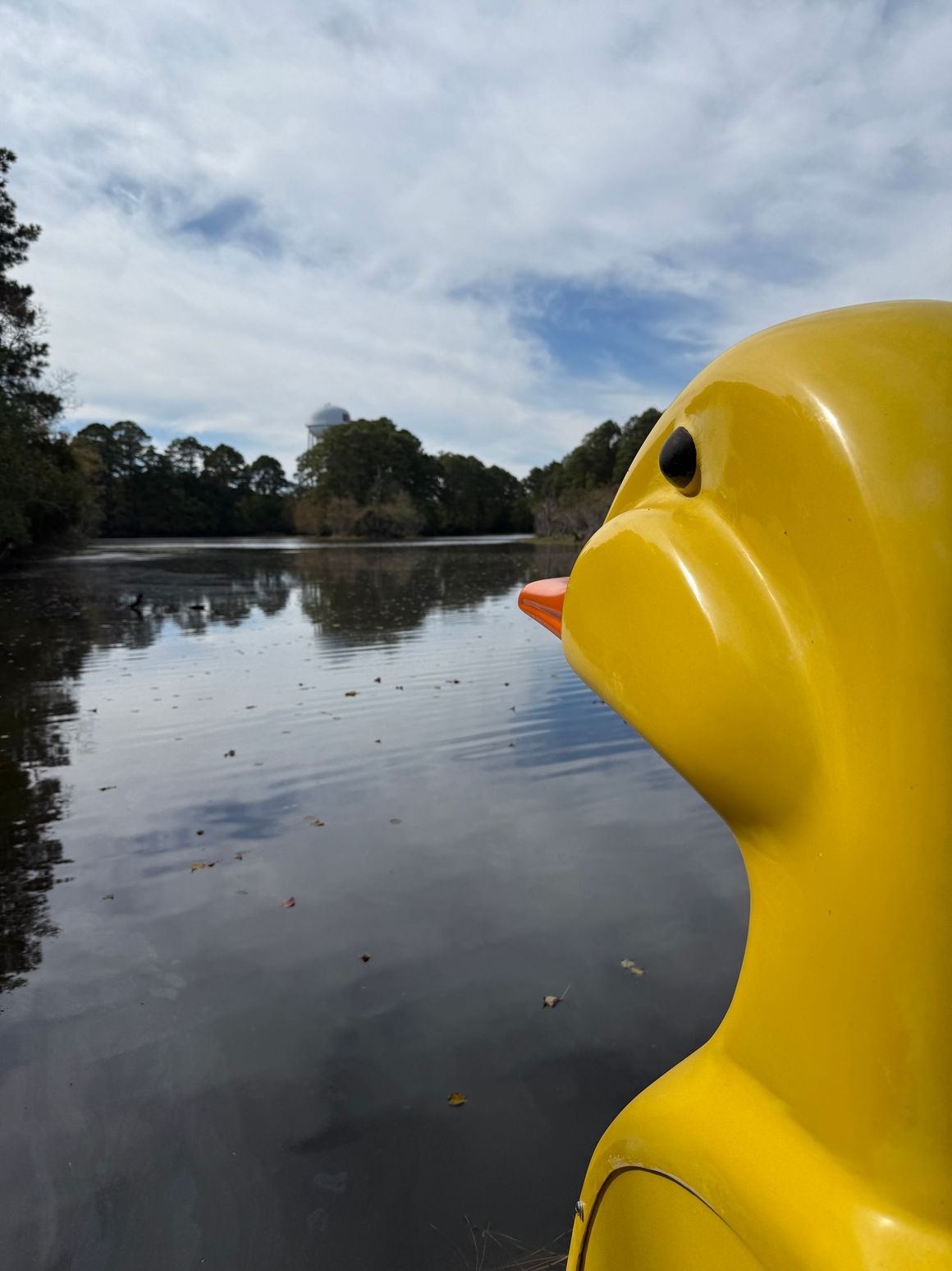 A large yellow duck sculpture looking out over a calm, tree-lined lake under a cloudy sky.