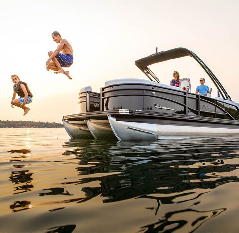 Two people jump from a black pontoon boat into calm water while others relax on board at sunset.