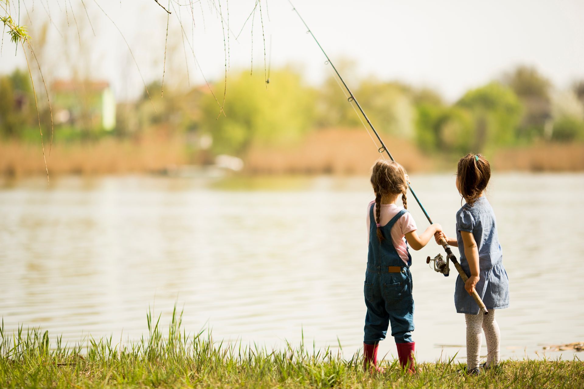 Two children in casual clothes stand by a calm lake, holding a fishing rod together while looking out at the water.