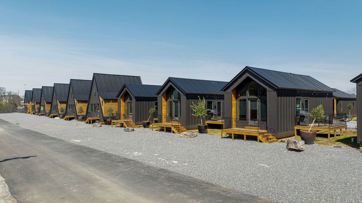 A row of modern, dark-colored small cabins with wooden accents and sloped roofs sitting on a gravel lot under a blue sky.