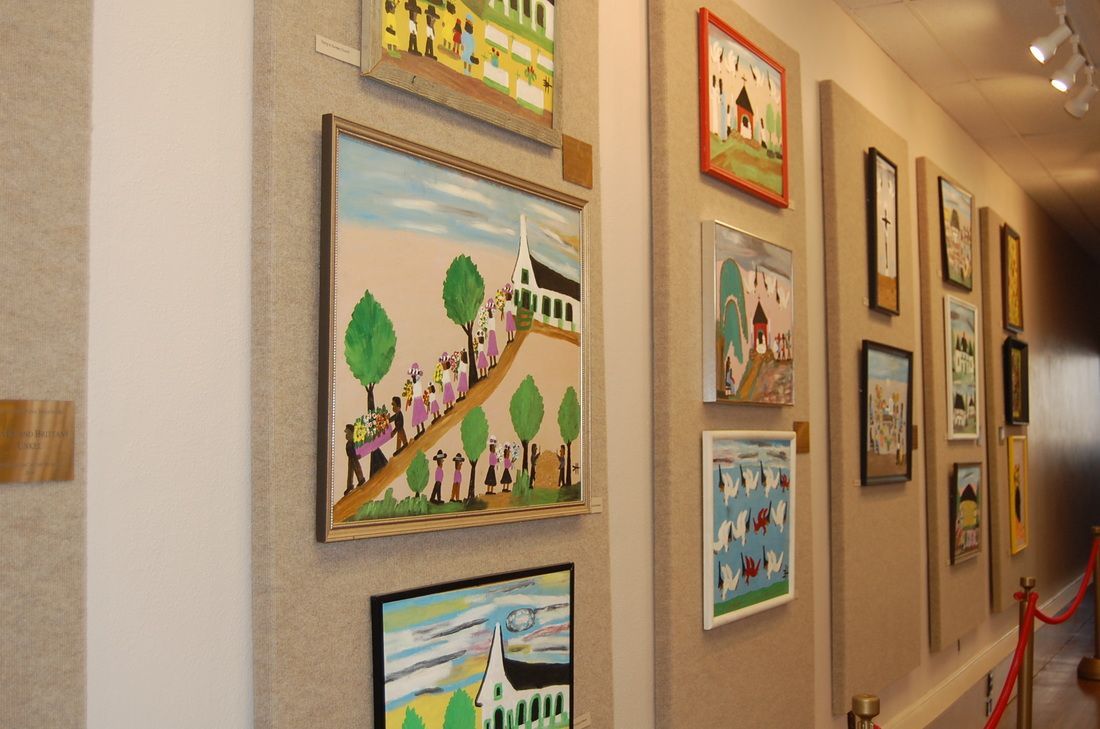 An art gallery hallway with framed folk-art style paintings displayed on tan fabric wall panels.