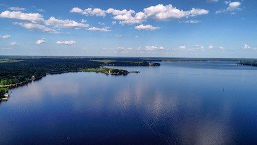 An aerial view of a wide blue lake under a bright sky with scattered white clouds, bordered by a dark green tree line.